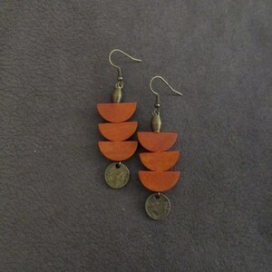 May include: A pair of dangle earrings featuring two stacked wooden semi-circles in a warm orange-brown tone, suspended from a bronze-colored metal hook with a small ribbed bead and a circular bronze-colored metal charm at the bottom of each piece.