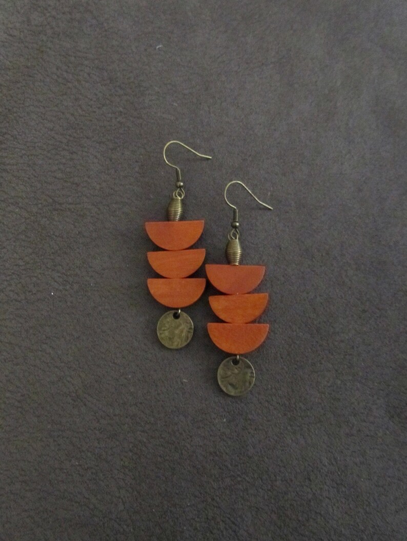 May include: A pair of dangle earrings featuring two stacked wooden semi-circles in a warm orange-brown tone, suspended from a bronze-colored metal hook with a small ribbed bead and a circular bronze-colored metal charm at the bottom of each piece.