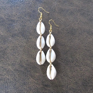May include: A pair of dangle earrings featuring three white cowrie shells per earring, strung together with gold-colored wire and hooks. The earrings are displayed against a dark brown background.