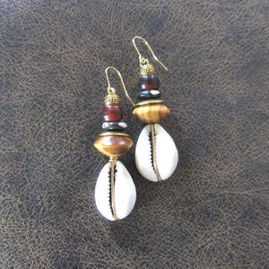 May include: A pair of handcrafted earrings featuring a combination of natural elements. The earrings showcase a white cowrie shell, wooden beads, and dark brown and red beads, all suspended from gold-colored ear wires. The design is a unique blend of textures and colors.