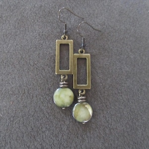 May include: A pair of dangle earrings featuring rectangular bronze-colored frames and round, pale green beads. The earrings have a simple hook design and are displayed against a gray background.