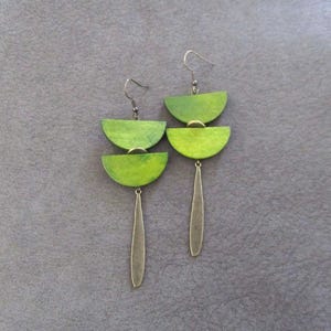 Puede incluir: Un par de pendientes colgantes con dos semicírculos de madera de color verde lima apilados, unidos por pequeños anillos de metal dorado y terminados con una pieza colgante de metal dorado, larga, delgada y texturizada en la base.