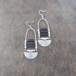 May include: A pair of dangle earrings featuring a silver-toned wire arch, three horizontal black rectangular beads, and a silver-toned semi-circular charm with concentric etched lines. The earrings are suspended from silver-toned metal hooks.