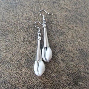 May include: A pair of dangle earrings featuring white seashells encased in silver-colored metal. The earrings have a long, conical design with a spiral wire detail and a hook closure. The background is a textured brown surface.