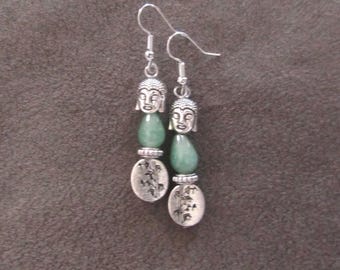 May include: Silver-toned dangle earrings featuring a Buddha head, a green teardrop-shaped bead, and an oval charm with a floral design. The earrings have a hook closure and are displayed against a brown background.