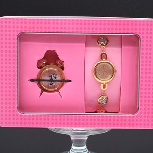 May include: A pink gift box with a clear window showing a gold-tone wristwatch and a gold-tone alarm clock. The wristwatch has a crystal-encrusted band.