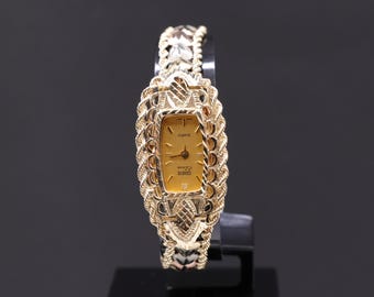 May include: A gold-tone wristwatch with a rectangular face and a gold-tone band. The watch face has a white background with black Roman numeral hour markers and black hands. The band is made of a braided design.