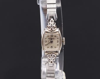 May include: A silver wristwatch with a white face and black Roman numeral hour markers. The watch has a silver band with a diamond-shaped design at the top.