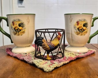 May include: Two cream-colored ceramic mugs with green handles and a rooster illustration, positioned on either side of a small, dark metal wire candle holder featuring a three-dimensional rooster figure attached to the front.