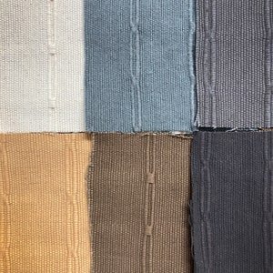 May include: A close-up of six fabric swatches in various colors. The colors include white, light blue, gray, tan, brown, and dark gray. Each swatch has a textured weave and a vertical seam down the center.