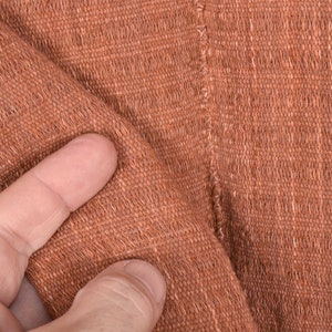 Peut inclure: Gros plan sur un tissu tiss&eacute; brun avec une surface textur&eacute;e. Le tissu est pli&eacute; et un doigt touche la surface.