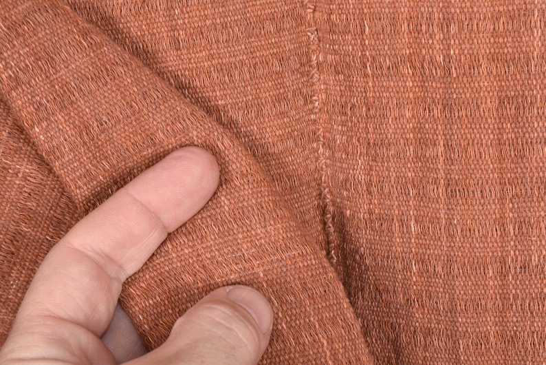 Peut inclure: Gros plan sur un tissu tiss&eacute; brun avec une surface textur&eacute;e. Le tissu est pli&eacute; et un doigt touche la surface.
