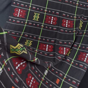 May include: Black fabric with a detailed pattern featuring red, white, yellow, and green geometric shapes. The design includes vertical and horizontal lines, creating a grid-like appearance. The fabric is folded, showing the intricate design.