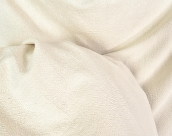 Könnte beinhalten: Nahaufnahme eines cremefarbenen Stoffes mit strukturierter Oberfläche. Der Stoff ist gefaltet und drapiert, wodurch weiche Schatten und Lichtreflexe entstehen. Das Material scheint eine Naturfaser zu sein, möglicherweise Leinen oder Baumwolle.