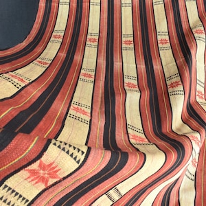 Puede incluir: Textil con un patrón repetitivo de rayas verticales en negro, rojo y beige. Las rayas se intercalan con motivos geométricos y florales. La tela tiene una textura ligeramente arrugada, lo que sugiere que está hecha de una fibra natural.