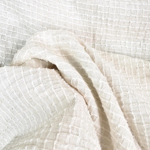 Puede incluir: Primer plano de una tela color crema con un patrón texturizado en forma de rejilla. La tela está drapeada, creando pliegues y sombras suaves. El material parece ligero y transpirable, adecuado para ropa o decoración del hogar.