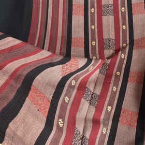 May include: A close-up of a woven fabric with a striped pattern in shades of red, black, and brown. The fabric has a subtle texture and a traditional design.