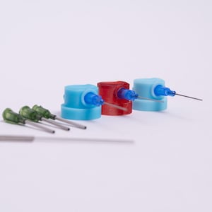 Pode incluir: Quatro tampas de agulha de plástico azul e vermelho com quatro agulhas prateadas sobre uma superfície branca.