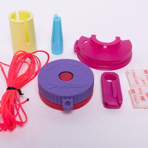 May include: A collection of colorful craft tools and accessories. Includes a purple and red design tool with the text "A Dulour Design Tool", a pink clip, a yellow cylinder, a blue cone, a pink lid, red string, and two adhesive strips.