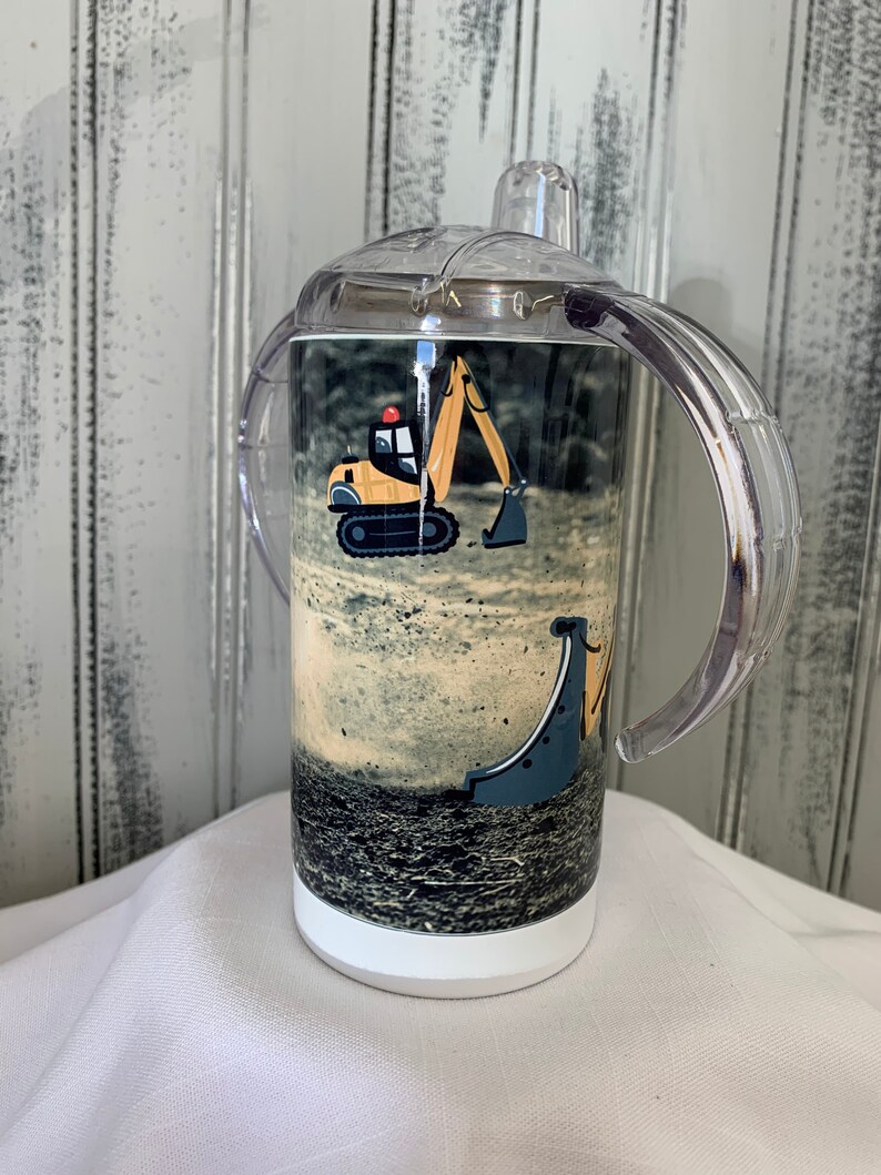 May include: A white stainless steel tumbler with a clear plastic lid and two handles. The tumbler has a printed image of a yellow toy excavator on a dirt background.