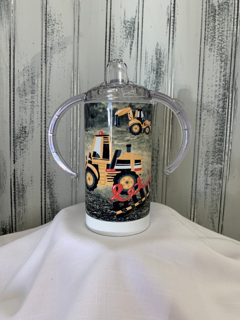 May include: A clear plastic sippy cup with two handles and a yellow and black construction vehicle design. The cup has a white lid with a clear spout.