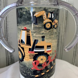 May include: A clear plastic sippy cup with two handles. The cup has a white base and a colourful design featuring yellow construction vehicles on a black and brown background.