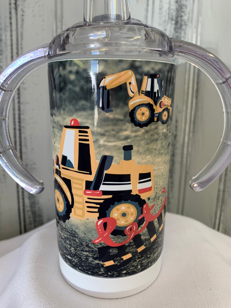 May include: A clear plastic sippy cup with two handles. The cup has a white base and a colourful design featuring yellow construction vehicles on a black and brown background.
