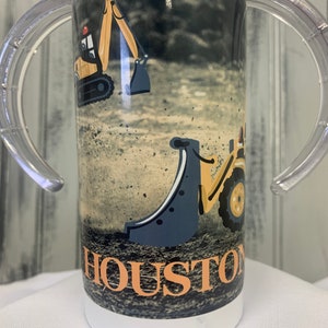 May include: A white tumbler with a black and yellow construction vehicle design. The tumbler has two clear plastic handles and the text "HOUSTON" is printed on the front.