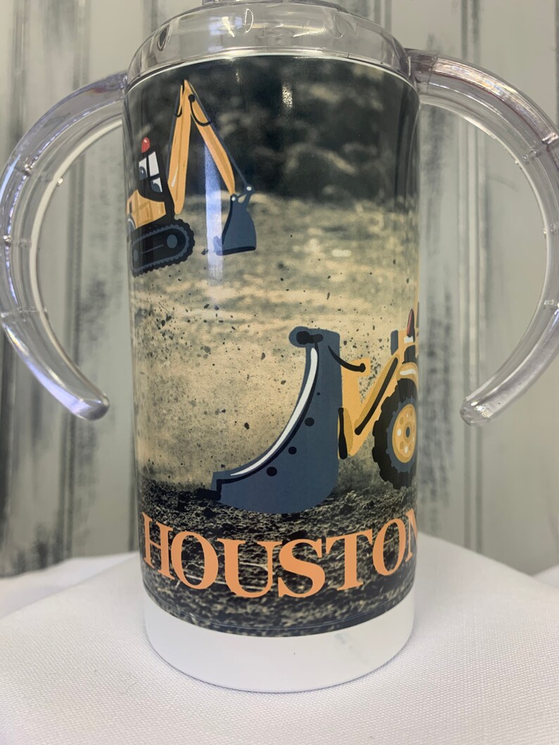 May include: A white tumbler with a black and yellow construction vehicle design. The tumbler has two clear plastic handles and the text "HOUSTON" is printed on the front.