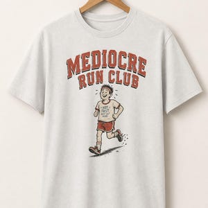 May include: A light gray short-sleeve t-shirt featuring a graphic of a runner with the text "MEDIOCRE RUN CLUB" in red and black letters. The runner wears a red headband, red shorts, and a shirt with the text "NOT FAST JUST OUT HERE".