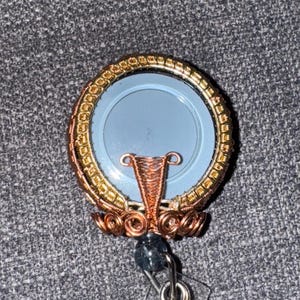May include: A handcrafted badge reel with a light blue center. The reel is encircled by gold and copper-colored wire, with intricate wirework details. A dark blue bead and a clear plastic clip complete the design.