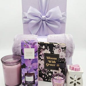 May include: A lavender gift set with a box tied with a large bow, a candle, a journal with the text "Bloom With Grace", a violet-scented bath product, a wax warmer, and a small candle.
