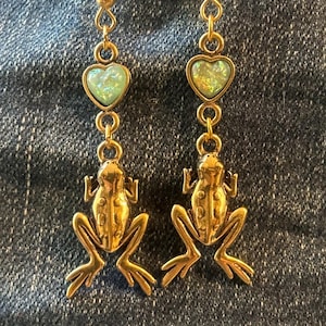 May include: A pair of gold-colored earrings featuring a frog design. Each earring has a heart-shaped, iridescent green stone above the frog. The earrings are hanging against a denim background.