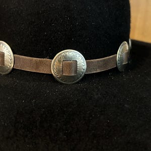 May include: Black hat with a brown leather band adorned with silver-colored, circular embellishments. The hat's brim is slightly curved, and the band features three decorative metal pieces.