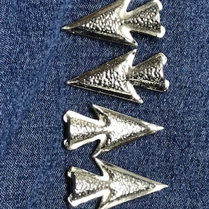 May include: Four silver-colored arrow-shaped embellishments with textured surfaces are arranged diagonally on a blue denim background. The arrowheads have a detailed, slightly granular texture, and the overall design is symmetrical.