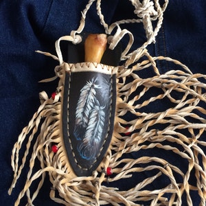 May include: A leather knife sheath with a fringe and red beads. The sheath is decorated with a painted design of feathers.