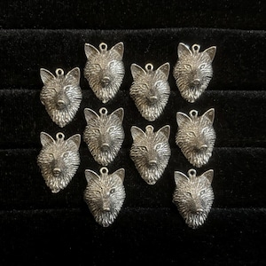May include: A collection of eleven silver-colored wolf head charms. Each charm features detailed facial features and a small loop at the top for attaching to jewelry or other crafts. The charms are arranged on a black surface.