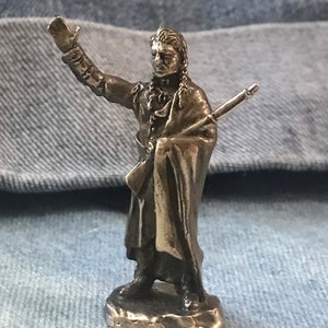 May include: A small, silver-colored figurine of a man in a long coat, holding a rifle and raising his right hand. The figure has long hair and is standing on a small, round base. The figurine is detailed and appears to be made of metal.