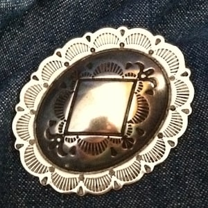 May include: A silver Navajo Concho with a diamond-shaped design in the center. The Concho has a scalloped edge and is decorated with intricate details.
