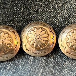 May include: Three circular silver-toned metal buttons featuring a central embossed starburst pattern surrounded by a series of small etched circles and radiating lines along the outer edge, arranged in a horizontal row on a dark blue denim fabric.