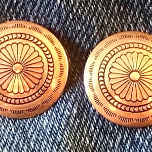 May include: A pair of round, copper-colored buttons with a decorative, floral design. The buttons have a raised, circular pattern with a smaller circle in the center.