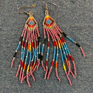 May include: A pair of beaded earrings with a triangular top and long fringe. The earrings feature a colorful design with pink, blue, red, yellow, and black beads. The earrings are on a denim background.