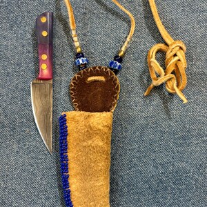 May include: A leather knife sheath with blue beads and a brown leather flap. The sheath is attached to a brown leather cord with blue and white beads. A small, patterned knife is next to the sheath.