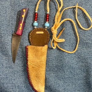 May include: A small, handmade knife with a brown leather sheath and a beaded lanyard. The knife has a dark metal blade and a red and purple handle with gold accents.
