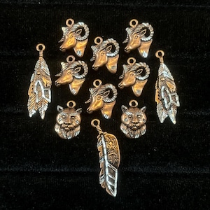 May include: A collection of bronze-toned metal charms, including ram heads, cat faces, and feather pendants. The charms have a vintage, antique appearance, with intricate detailing and small loops for attaching to jewelry or other crafts.