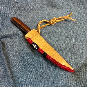 May include: A small knife with a dark wooden handle is sheathed in a tan leather case. The case is decorated with red, black, and white beaded accents. A tan leather cord is tied at the top of the sheath.
