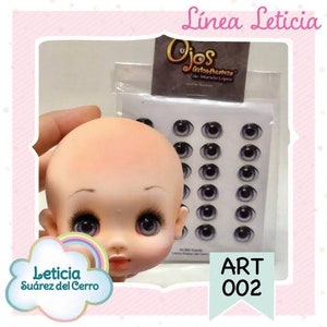 May include: A porcelain doll head with painted eyes and a rainbow cloud behind it. The doll head is part of a craft kit for making dolls. The kit includes a sheet of self-adhesive doll eyes with a black and white iris and a blue pupil. The sheet of eyes is labeled "Ojos" and "Art 002".