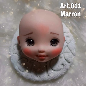 May include: A porcelain doll head with painted brown eyes, rosy cheeks, and a closed-mouth smile. The head is on a white base with the word "CAPRICORN" written on it. The text "Art.011 Marron" is in the upper right corner of the image.