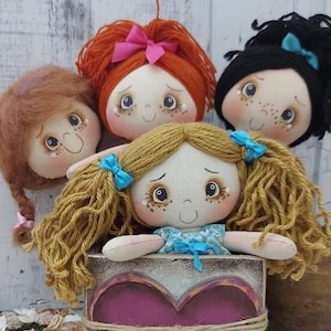 Op de afbeelding: Vier handgemaakte stoffen poppen met garenhaar, zittend in een houten doos met een hartvormige uitsparing. De poppen hebben geschilderde gezichten met roze wangen en grote ogen. De doos is wit geverfd met een roze hart en een bruine touw strik.