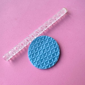 May include: A clear acrylic rolling pin with a repeating floral petal pattern and a matching circular blue clay disc featuring the same embossed geometric flower design. The tools are designed for creating textured impressions in soft materials.